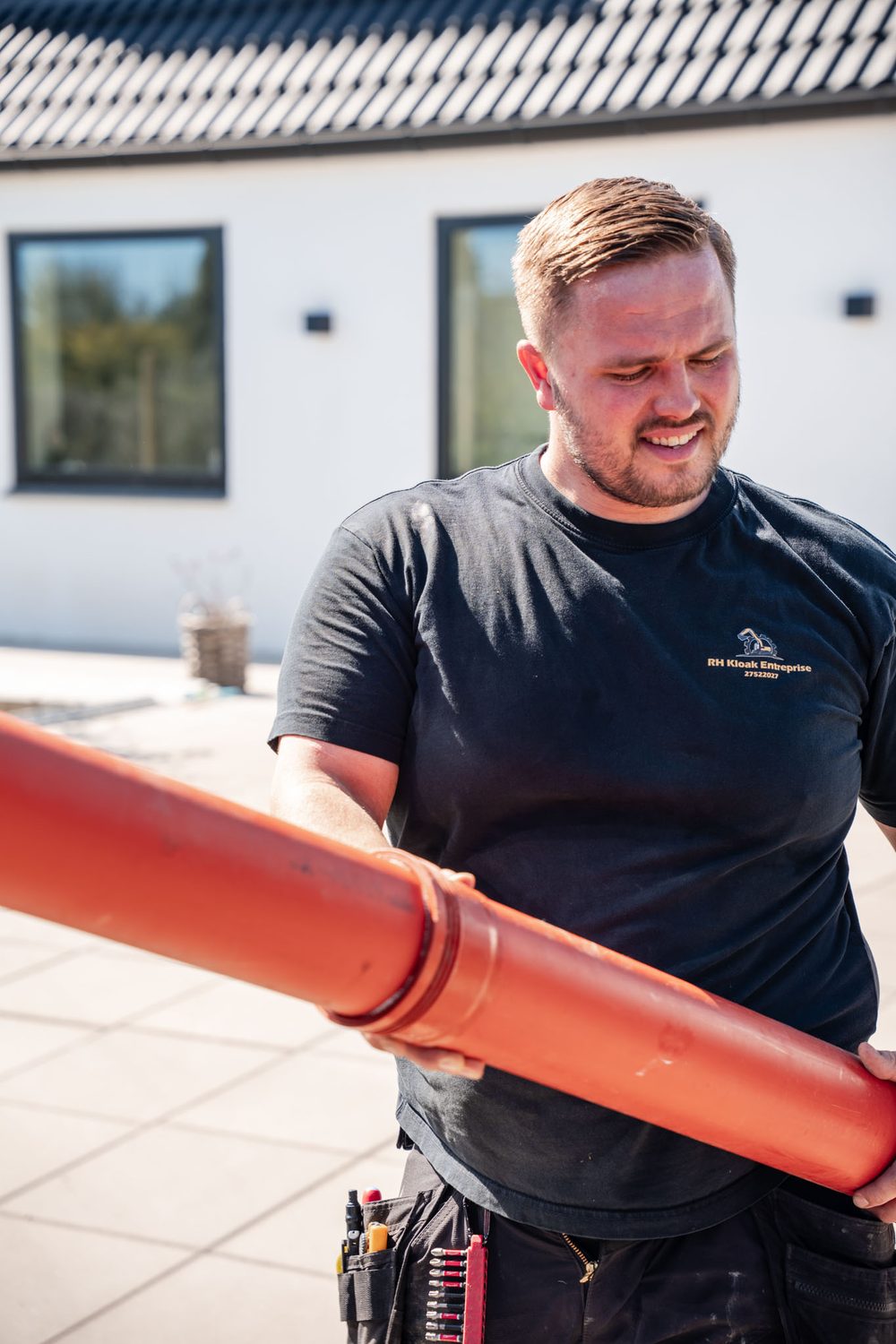 Rasmus bærer et rødt kloakrør foran et hvidt hus